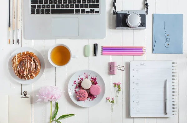 Жіночий плоский робочий простір з ноутбуком, чашкою чаю, макаронами, фотокамерою та квітами на білому дерев'яному столі. Вид зверху сміється . — стокове фото