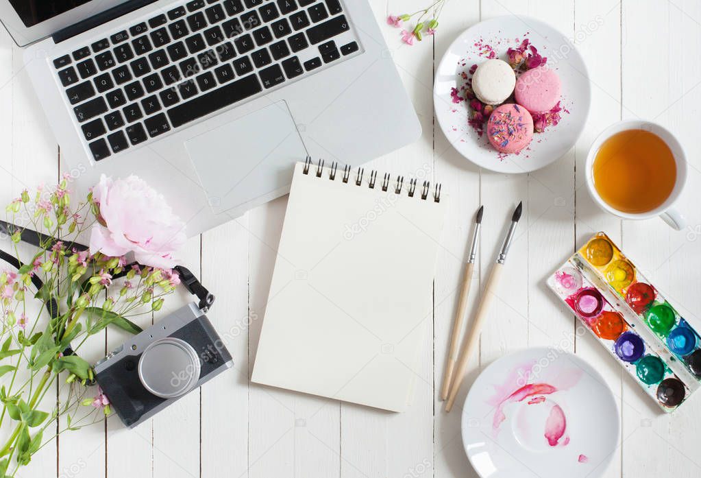Feminine flat lay workspace with watercolor, paintbrushes, laptop, cup of tea, photo camera and flowers on white wooden table. Top view mock up.