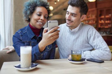 Afrikalı kadın ve çay ve kahve içme kafede oturan beyaz adam genç çift portre. Erkek bir şey ona bakıyorsun adam için smartphone gösteriyor, onlar her ikisi de gülüyorsun.