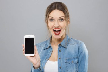 Şık güzel genç kadın boş cep telefonu sunulması ve şaşkınlık ile açık ağız ile gülümseyen gri arka plan üzerinde izole kapalı bir kadeh. Boşaltmak için reklam mal ve hizmet.