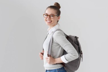 Rahat kıyafetler ve modaya uygun gözlük ile sırt çantası sırtında sanki boş zaman, tatil ve toplantılar tahmin gülümseyen izole üzerinde gri arka plan ayakta mutlu genç kadın portresi.