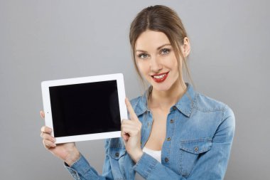Portre çekici genç kız yavaşça tablet bilgisayarın boş ekran gösterilen süre gülümseyen gri arka plan üzerinde izole kırmızı ruj ile kayıtsızca giyinmiş. Boşaltmak için sunulan reklam.