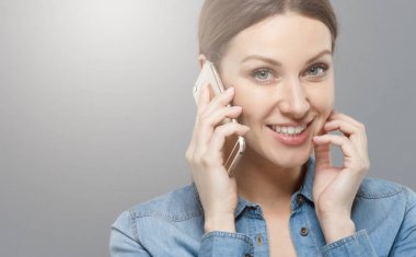 Portre fotoğraf kamera ve gülümseyen dostu düz seyir onun arkadaşı ile telefon görüşmesi sırasında yanağı parmaklarıyla dokunarak gri arka plan üzerinde izole çekici genç kadın.
