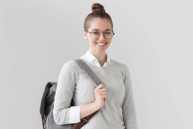 Gri trendy sırt çantası giyiyor ve ileri kayış, doğruca kameraya, seyir gülümseyen ve iş ya da eğlence zaman tahmin çekerek gri arka plan üzerinde izole genç Avrupa kadın fotoğrafı.
