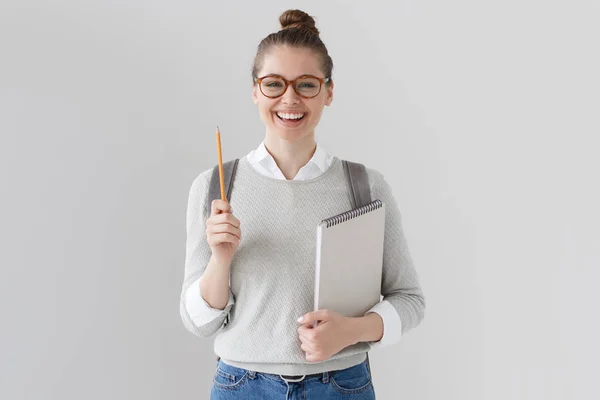 积极的学生女孩上灰色的背景，戴着眼镜、 毛衣和牛仔裤，孤立的影棚拍摄背包，向上与铅笔，拿着笔记本，看起来兴奋、 充满活力和想法. — 图库照片