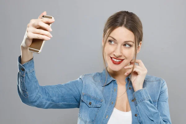 Foto de mulher atraente com maquiagem isolada em fundo cinza tocando sua bochecha com os dedos e esticando o braço com smartphone para tirar foto de selfie para compartilhá-la em redes sociais . — Fotografia de Stock