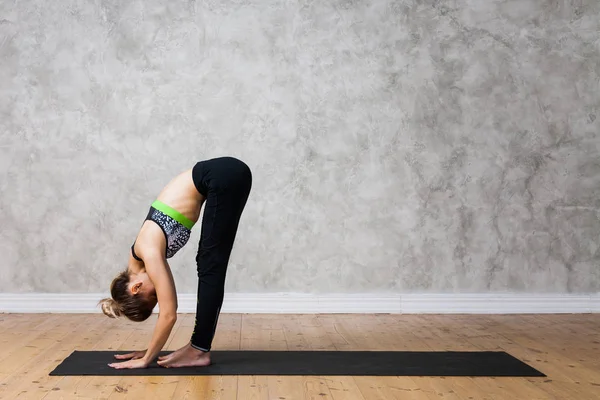 Young woman practicing Standing forward bend, Uttanasana yoga pose against texturized wall / urban background