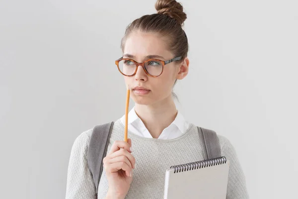 Nahaufnahme Porträt einer gut aussehenden jungen Frau, die isoliert auf grauem Hintergrund steht, eine Brille trägt, den Bleistift neben die Lippen hält, nach links denkt und sucht, um Antwort oder Lösung zu finden.. — Stockfoto