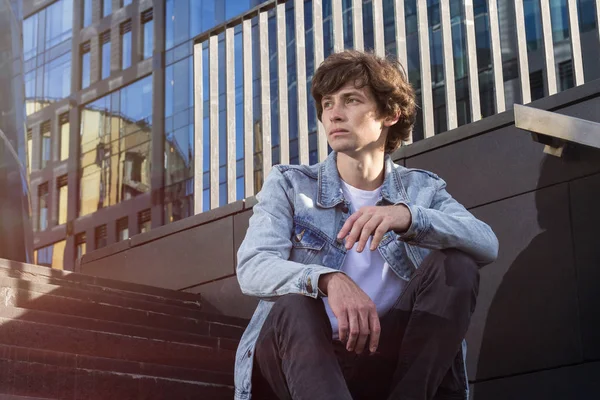 Portret przystojny zadumany młodzieniec noszenie Kurtki jeansowe, siedząc na schodach na tle miasta — Zdjęcie stockowe