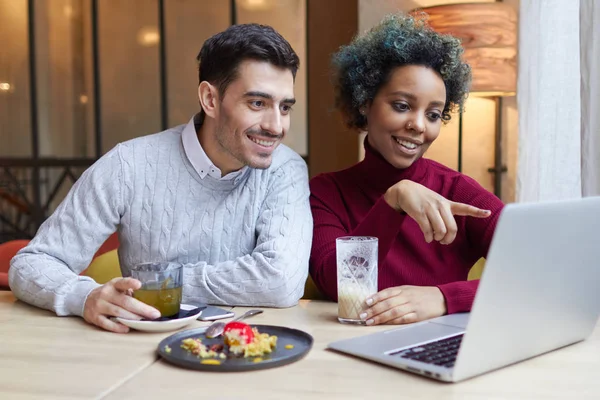 Portret van gelukkig liefhebbers ontmoeting binnenshuis met laptop open op tafel in café. Donkere meisje wijst om weer te geven met vrolijke glimlach, man kijkt aandachtig scherm betrokken bij wat ze toont. — Stockfoto