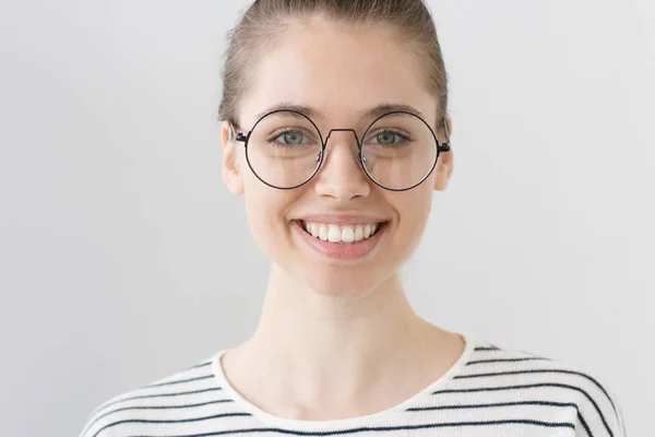 Primer plano de la joven adolescente europea aislada sobre fondo gris llevando gafas con borde negro delgado, sonriendo al fotógrafo, luciendo feliz, positiva y optimista, abierta a la comunicación . — Foto de Stock