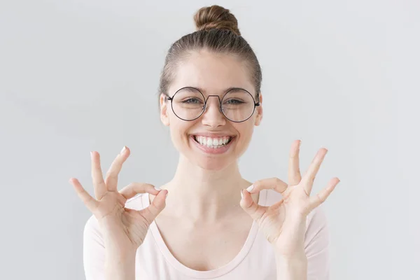 Inomhus bild av vacker flicka isolerade på grå bakgrund bär stora runda glasögon och ser glad och positiv som hon visar dubbla ok tecken, att vara säker på bästa resultat i framtiden. — Stockfoto