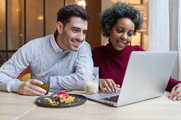 Nahaufnahme von jungen interrassischen Liebhabern, die Zeit im Café verbringen und gemeinsam am Laptop die Medien beobachten. Afrikanische Amerikanerin benutzt Touchpad, kaukasischer Mann schaut auf Display mit Überraschung hält Teetasse. — Stockfoto