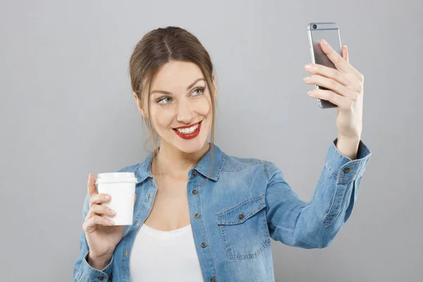 Close-up de jovem mulher bonita vestindo batom vermelho, isolado no fundo cinza, virou à direita com o braço segurando celular, tirando foto selfie e segurando tomar café em outra mão, sorrindo . — Fotografia de Stock