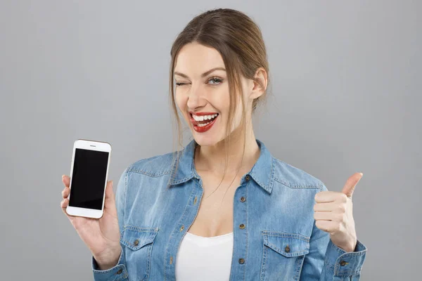 Retrato interno de menina europeia atraente isolado em fundo cinza vestindo maquiagem, segurando smartphone em branco, piscando com um olho e mostrando gesto de aprovação, sentindo-se otimista e feliz . — Fotografia de Stock