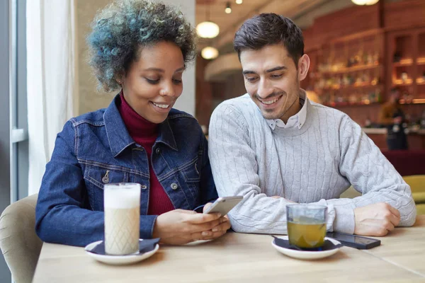 Plan intérieur d'une fille à la peau foncée et d'un mec européen passant un agréable moment dans un café. Fille avec les cheveux teints en bleu avec smartphone profite wi-fi gratuit, gars regarde de côté sur ce qu'elle parcourt . — Photo