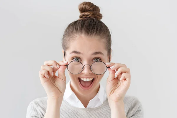 Retrato interno de mulher europeia excitada com cabelo amarrado em coque isolado em fundo cinza com óculos de armação preta elegantes, sua boca aberta de surpresa com notícias positivas ou boa oferta . — Fotografia de Stock