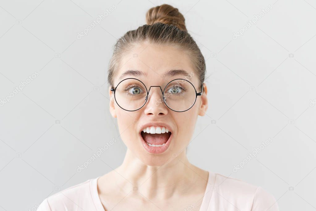 Portrait of positive European girl in light pink top isolated on grey background wearing thin-rimmed eyeglasses, unable to believe in news concerning surprising details of what she is looking at.