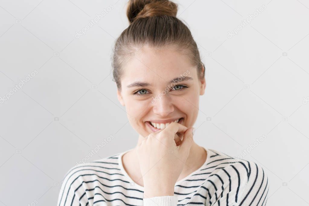 Beautiful green-eyed European female in casual clothes looking at camera, smiling, touching mouth with fingers in expression of shyness, listening to interlocutor, looking happy and interested.