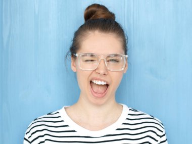 Gülümseyen ve göz kırpıyor, yüksek sesle bağırarak, mavi arka plan üzerinde duran bir mutlu genç yenilikçi Avrupa teen Kız bardaklarda kameraya oynak bir şekilde yanıp sönen nerdy kare portresi kapatın