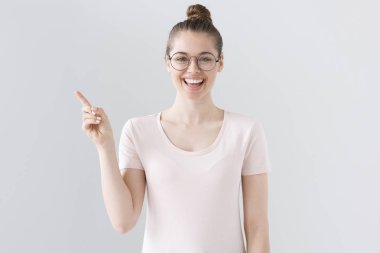 Olumlu gülümseyen bir el ve Dostu ile sol tarafı işaret eden genç çekici genç kız moda gözlük rahat kıyafetler ve saç modeli, gri arka plan üzerinde izole closeup çekim.