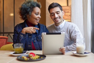 Pozitif genç ırklararası çift birlikte masada oturan ve tablet kullanarak zevk küçük restoranda fotoğrafı. Kız onun erkek resim çizim ekrana işaret ediyor. 