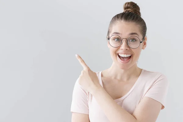 Övergripande bild av ung snygg kvinna isolerad på grå bakgrund bär stora runda Glasögon, pekar Vänsterriktad med pekfingret, ser oerhört glada, nöjda och förvånade. — Stockfoto