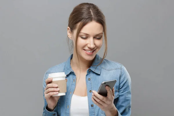 Close-up de jovem bela senhora europeia isolada no fundo cinza segurando copo de café de papelão e smartphone em suas mãos olhando para a tela e sorrindo alegremente para o conteúdo que ela está assistindo . — Fotografia de Stock