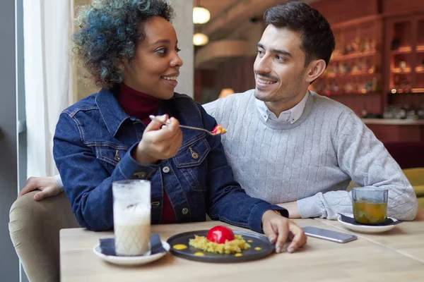 Jolie fille afro-américaine et mec caucasien manger dans un café. Femme tient cuillère avec morceau de dessert prêt à nourrir son amant, à la fois en se regardant avec soin et amour . — Photo