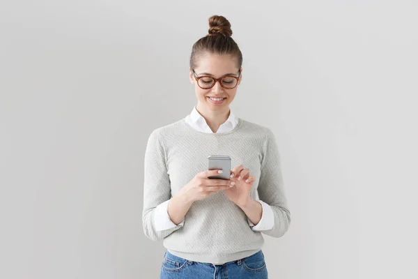 Foto interna de jovem atraente mulher europeia em pulôver cinza, camisa branca e óculos de jantes marrom isolados em fundo cinza com smartphone na mão, tela sensível ao toque com o dedo para digitar a mensagem . — Fotografia de Stock