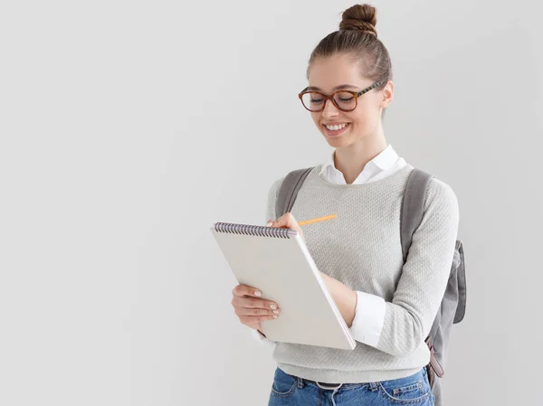 Foto interna de jovem mulher europeia no lado direito da foto contra fundo cinza tomando notas em folhas em branco de escrita pad ela está segurando com sorriso satisfeito e interessado, feliz em estudar . — Fotografia de Stock