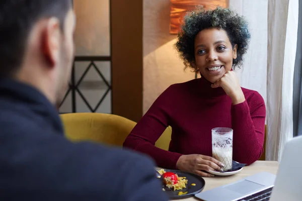 Indoor photo of smiling young dark-skinned woman drinking coffee in cafe and having dessert with laptop on table. Female is on date with European man in dark shirt, his back turned to viewer. — Stock Photo, Image