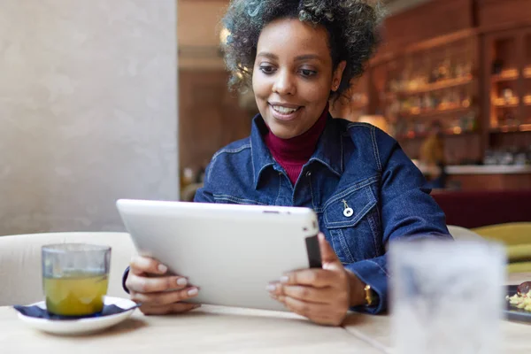 Attraktive ethnische Dame in blauer Jeansjacke sitzt am Cafétisch bei Tageslicht, Teetasse in der Nähe und hält ihren Tablet-PC glücklich lächelnd über positive Nachrichten und gute Bilder aus dem Web mit Inhalten. — Stockfoto