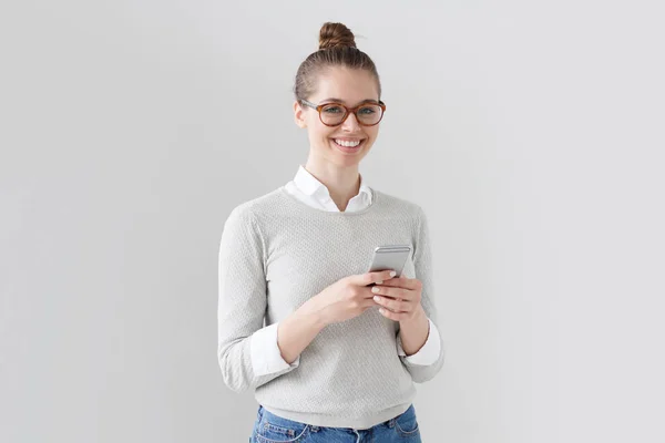 Porträtt av snygg college student tjej isolerad på grå bakgrund, tittar höger kameran medan du håller telefonen, skriva meddelanden och surfa Internet, leende med glädje från kommunikation. — Stockfoto