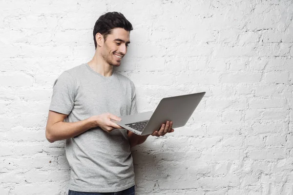 Retrato interior del hombre caucásico vestido con camiseta y jeans de pie contra la pared de ladrillo blanco sosteniendo la computadora portátil y viendo los medios con sonrisa feliz, compartiendo contenido web con compañeros o colegas . —  Fotos de Stock