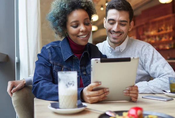 快乐的非洲裔美国女性特写欧洲白种人的男人坐在窗口，旁边的咖啡馆一起看显示的快乐与幸福的微笑的平板电脑上的媒体内容. — 图库照片