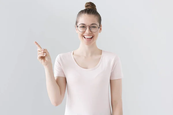 Close-up tiro de jovem atraente adolescente em óculos da moda isolado em fundo cinza com roupas casuais e penteado, apontando para o lado esquerdo com uma mão, sorrindo positivamente e amigável . — Fotografia de Stock