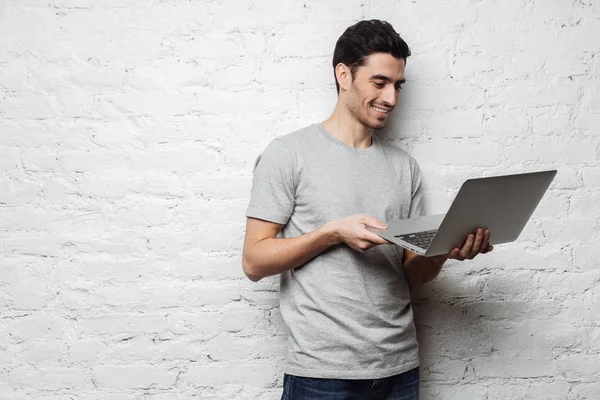 Estudio primer plano de hombre caucásico sonriente vestido con camiseta gris y jeans, de pie y sosteniendo el ordenador portátil en sus manos, navegar por páginas web, mirar a través de las fotos y comunicarse en línea con el placer . —  Fotos de Stock