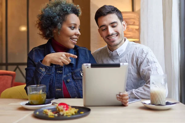 Photo d'un jeune couple interracial positif dans un petit restaurant assis à table ensemble et profitant de l'utilisation de la tablette. La fille pointe vers l'écran, attirant l'attention de ses petits amis sur l'image . — Photo