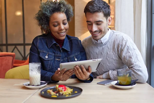 室内特写的年轻人微笑好看的非洲裔美国女孩和高加索的家伙穿得随便使用平板电脑在咖啡馆里一起浏览互联网，享受无线连接. — 图库照片