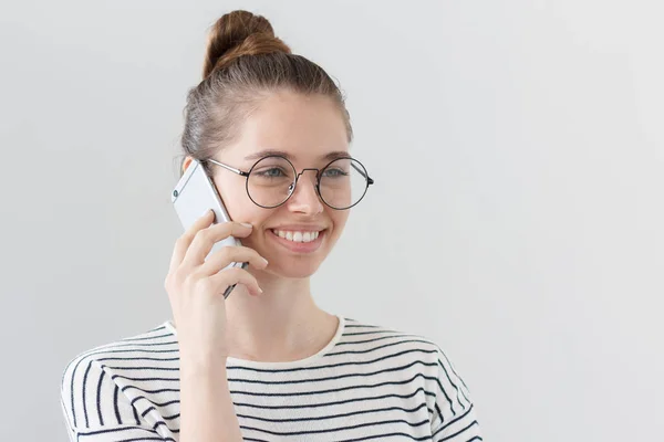 Sida porträtt av unga europeiska kvinnliga isolerade på grå bakgrund bär hipster Glasögon, lyssnar på samtalspartner på smartphone, visar nöjd lyckligt leende från nyheter hon hörs. — Stockfoto