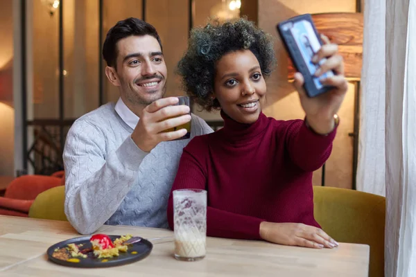 快乐异族情侣在下午在咖啡厅约会，深色皮肤的女人伸展她手捧着手机采取她和男友的自拍照，都是积极微笑和显示内容. — 图库照片