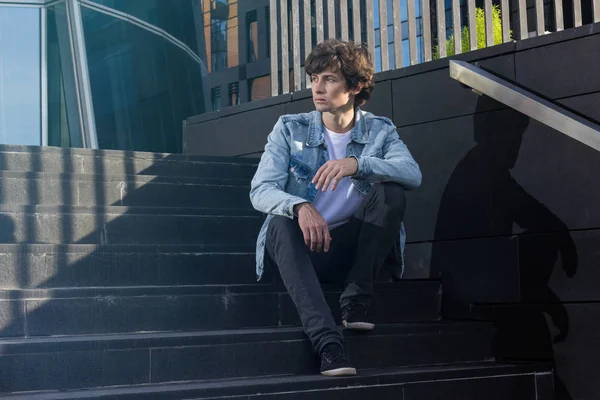 Kot ceket şehir arka plan karşı merdivenlerde oturan bir düşünceli yakışıklı genç portresi