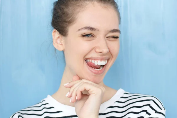 Bonito retrato de cerca de una joven europea. Feliz mujer linda engañosa con la cara sonriente parpadeando a la cámara de una manera lúdica, de pie sobre el fondo azul. Concepto de expresión de guiño — Foto de Stock