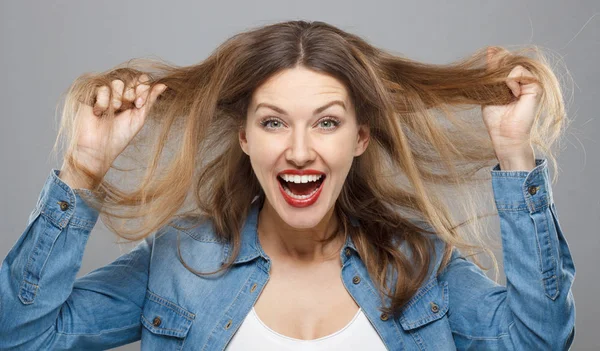 Closeup of cheerful emotional woman with makeup isolated on gray background showing wide open smile, stretching her hair upwards and screaming with happiness and delight.