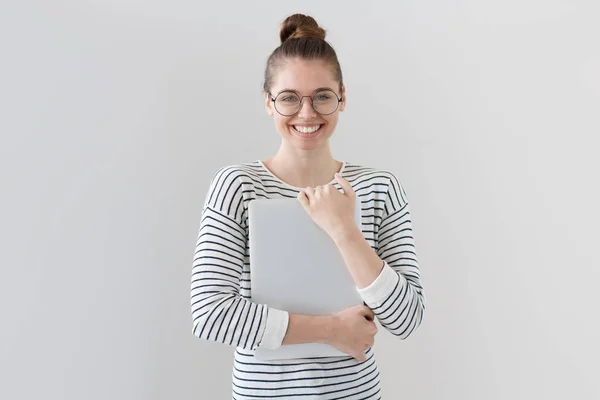 Close-up de jovem mulher europeia isolada em fundo cinza no topo listrado casual, segurando laptop nas mãos e pressionando-o para o peito com sorriso feliz e expressão de prazer e satisfação . — Fotografia de Stock