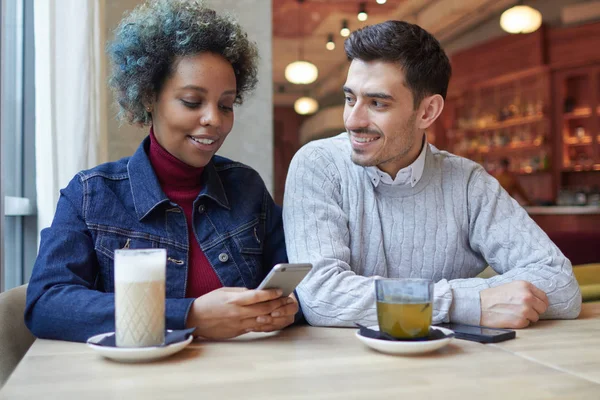 Attractive dark skinned woman spending time together with her European boyfriend in cozy cafe, holding cellphone, texting and browsing pages, her lover watching her with tenderness and love. — Stock Photo, Image