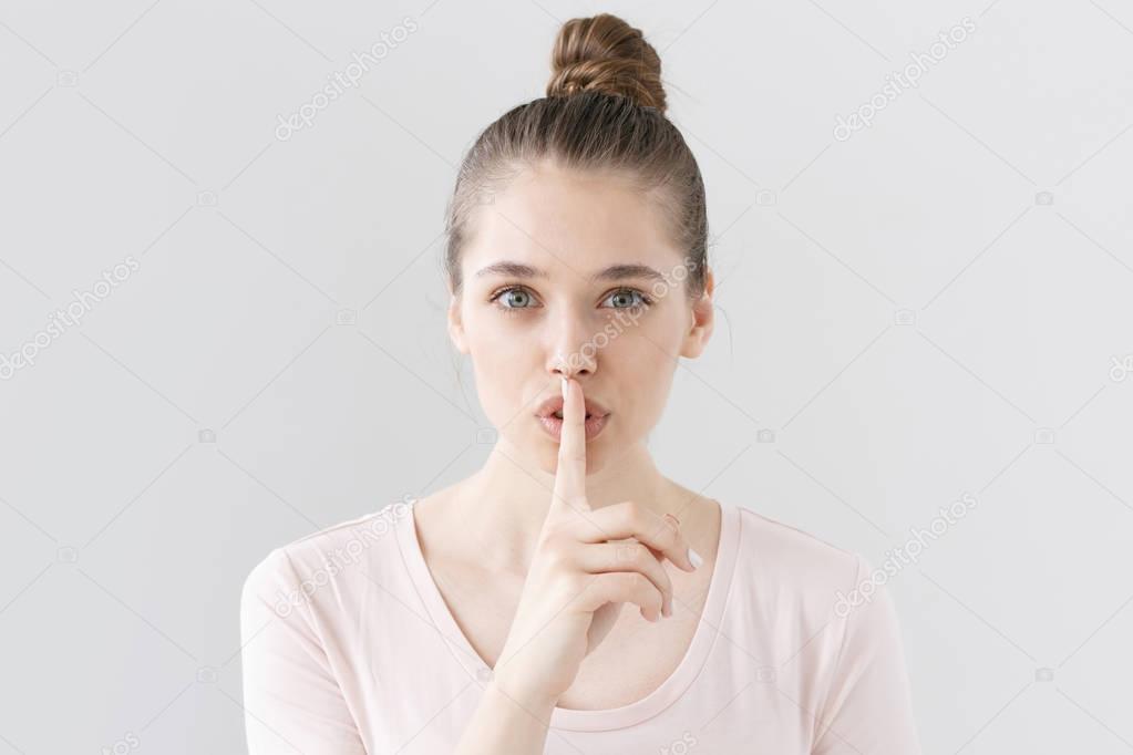 Horizontal picture of good-looking young woman isolated on grey background making gesture of silence in order to draw attention to important piece of news or secret that should not be disclosed.