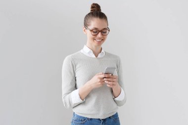 Elinde cep telefonu ile ayakta, faiz ve sıcak iyimser gülümseme ile ekrana bakarak, online iletişim kurmak için dört gözle gri arka plan üzerinde izole genç güzel kadın fotoğrafı.