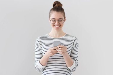 Gri arka planda izole edilmiş, rahat giyinmiş Avrupalı bir kızın cep telefonunun ekranına dikkatlice bakarken, web sayfalarına bakarken ve arkadaşlarıyla sohbet ederken gülümsediği yakın plan fotoğrafı..
